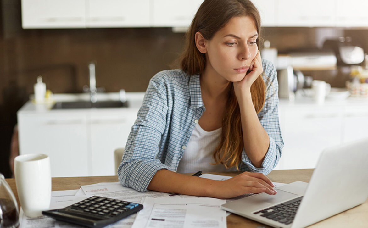 People, accounting, finances, family budget and financial issues concept. Serious young European woman calculating domestic expenses, sitting at dining table in front of open laptop computer