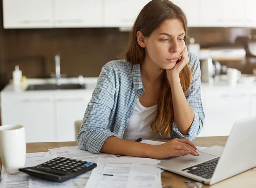 People, accounting, finances, family budget and financial issues concept. Serious young European woman calculating domestic expenses, sitting at dining table in front of open laptop computer