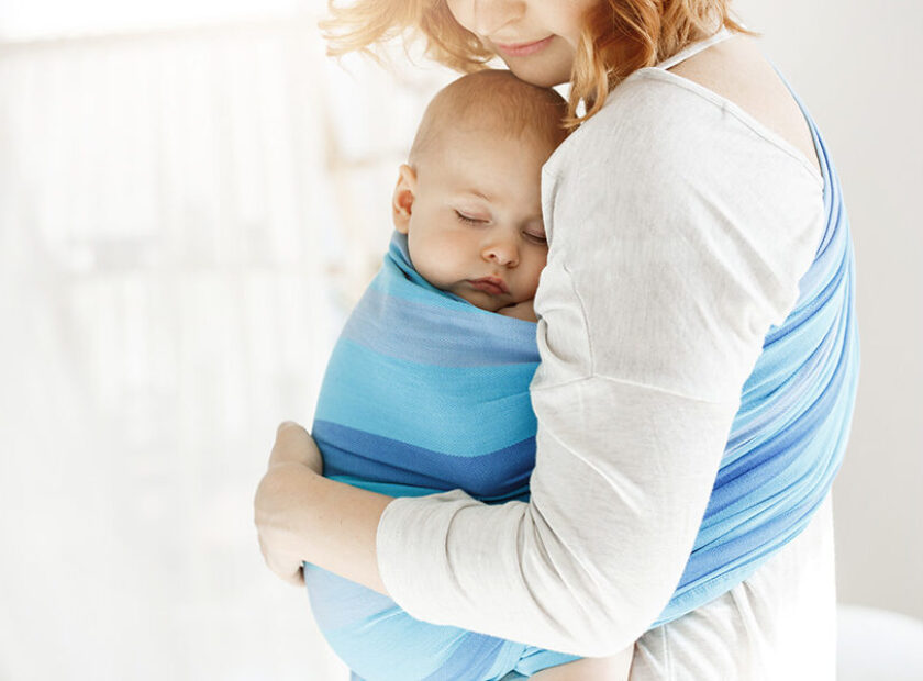 Tiny newborn child close eyes and having good sleep in baby sling feeling protection from his beautiful young mother. Family, lifestyle concept.