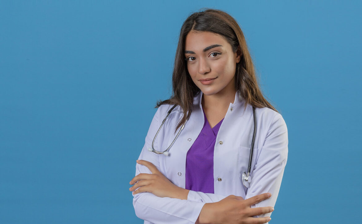 young woman doctor in white coat with phonendoscope looking confident standing with crossed arms over blue isolated background