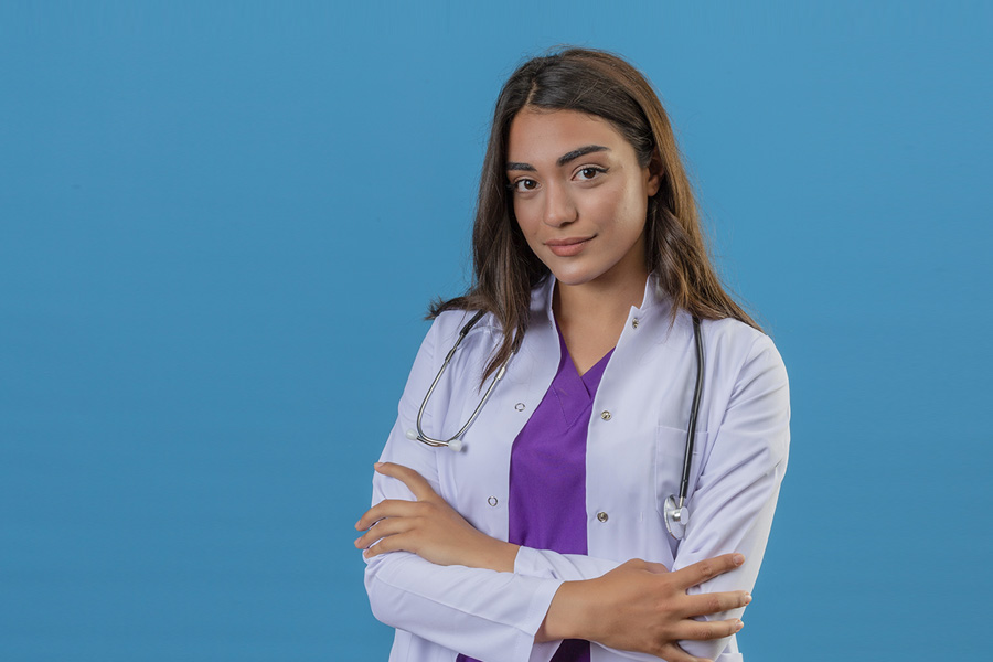 young woman doctor in white coat with phonendoscope looking confident standing with crossed arms over blue isolated background
