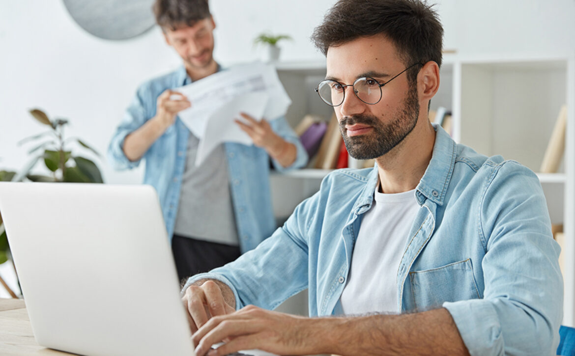 Business and startup concept. Bearded male freelancer works on laptop computer, keyboards information, thinks about profits, his partner looks attentively through papers, pose in spacious office