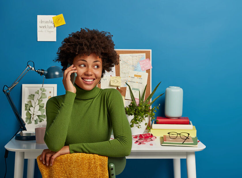 Afro American woman talks via smart phone, works at desk in home office, has cheerful face expression, dressed in casual wear, poses against desktop, isolated over blue wall with sticky notes