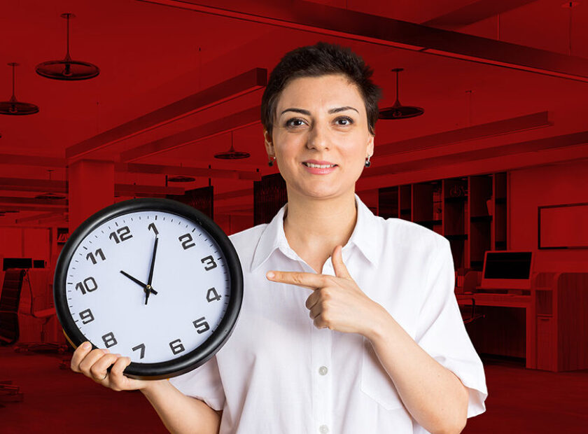 smiling middle-aged woman holding and pointing at clock looking at camera isolated on orange background