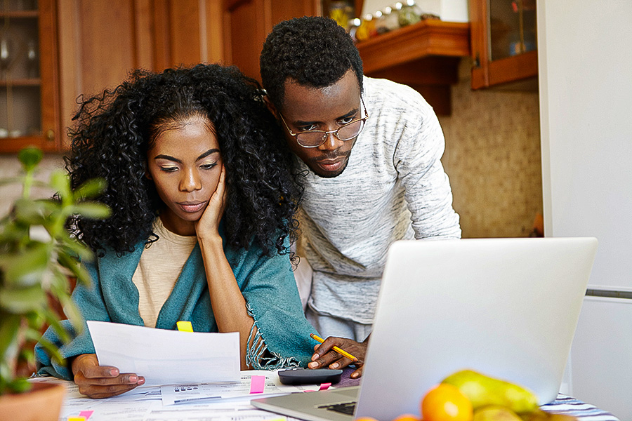 Young African family paying utility bills online using laptop computer. Unhappy female sitting at table analyzing sheet of paper in her hand, calculating domestic expenses together with her husband