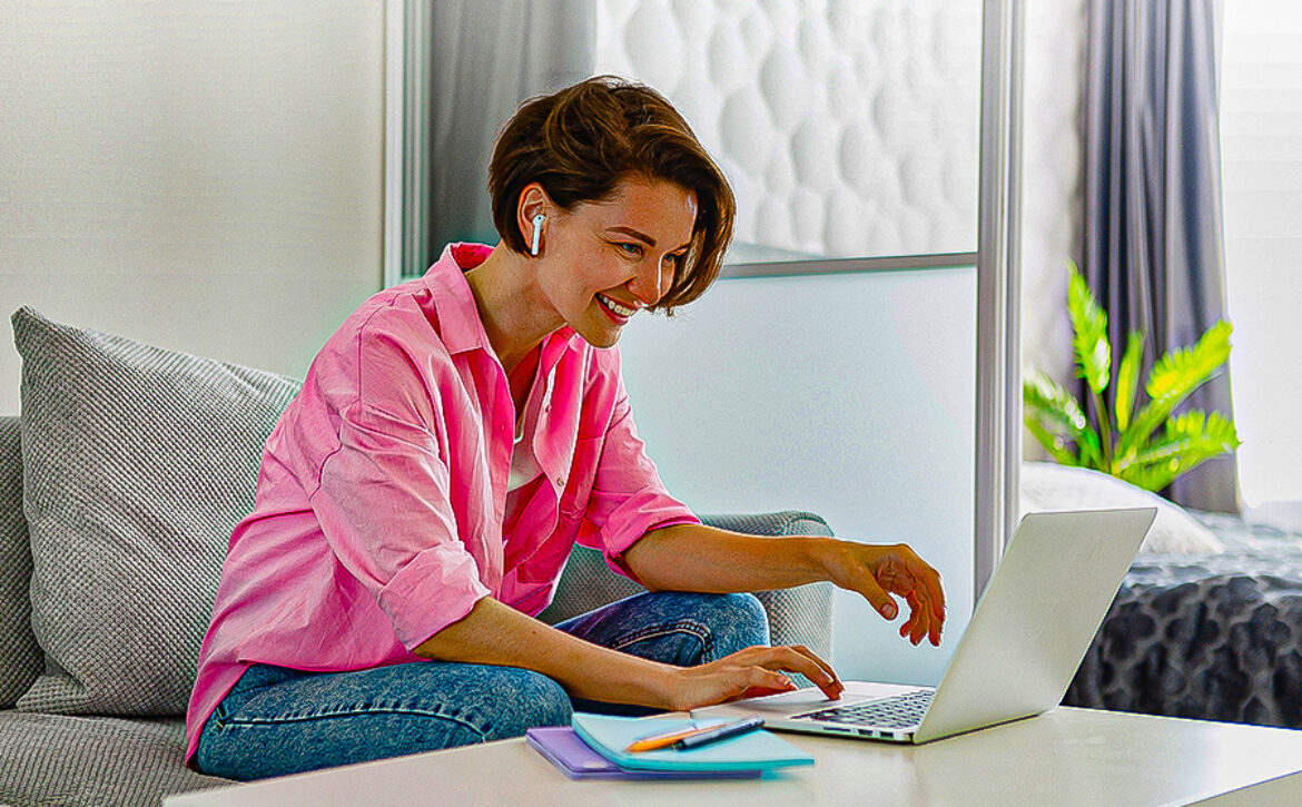attractive smiling woman in pink shirt sitting relaxed on sofa at home at table working online