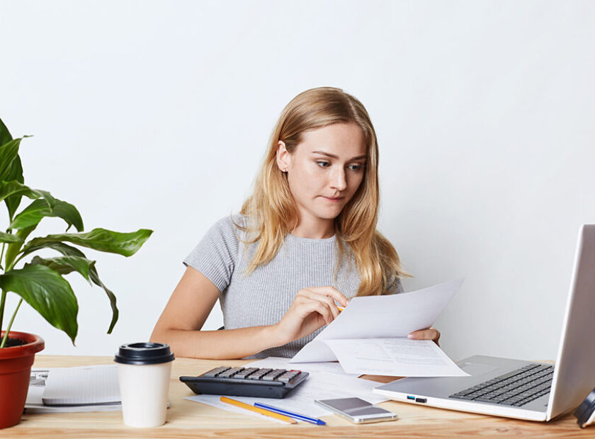 Portrait of young businesswoman working with laptop and calculator, looking attentively at documents, calcualting bills of company, making financial report. People, career and business concept